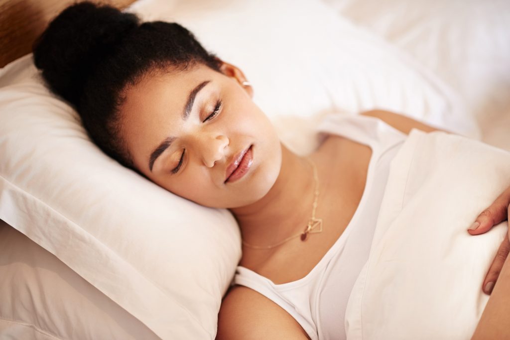 A woman with dark hair in a bun lies on her back in bed, eyes closed, resting her head on a white pillow and covered by a white blanket, enjoying restful sleep that supports her overall health and wellbeing.