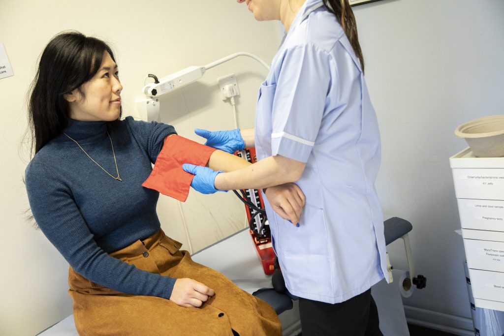 A healthcare worker measures a seated woman's blood pressure using a cuff in a medical examination room, highlighting the importance of regular consultations for preventative health.
