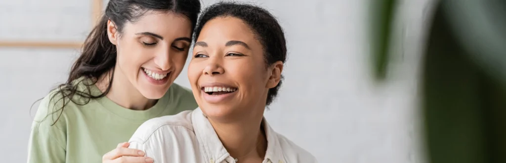 Two women smiling together, one standing behind the other with her hand on her shoulder, in a bright indoor setting.